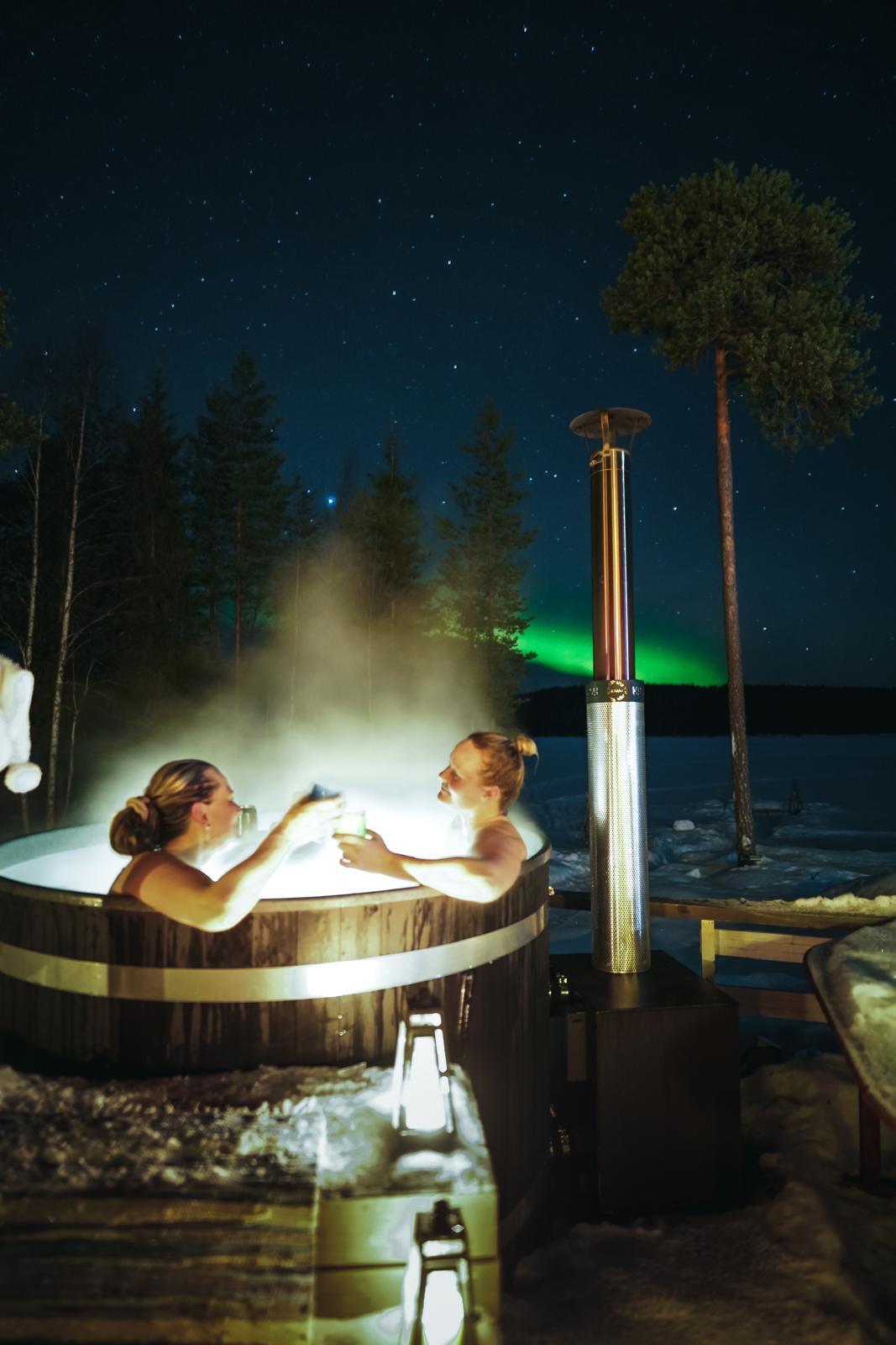 two women in a hot tub with green northern lights in the background. Lapland Royal Adventures, Rovaniemi, Lapland, Finland