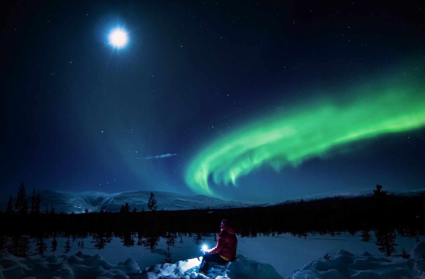 Person sitting in snow watching green northern lights in the sky. Tour by LaplandScapes