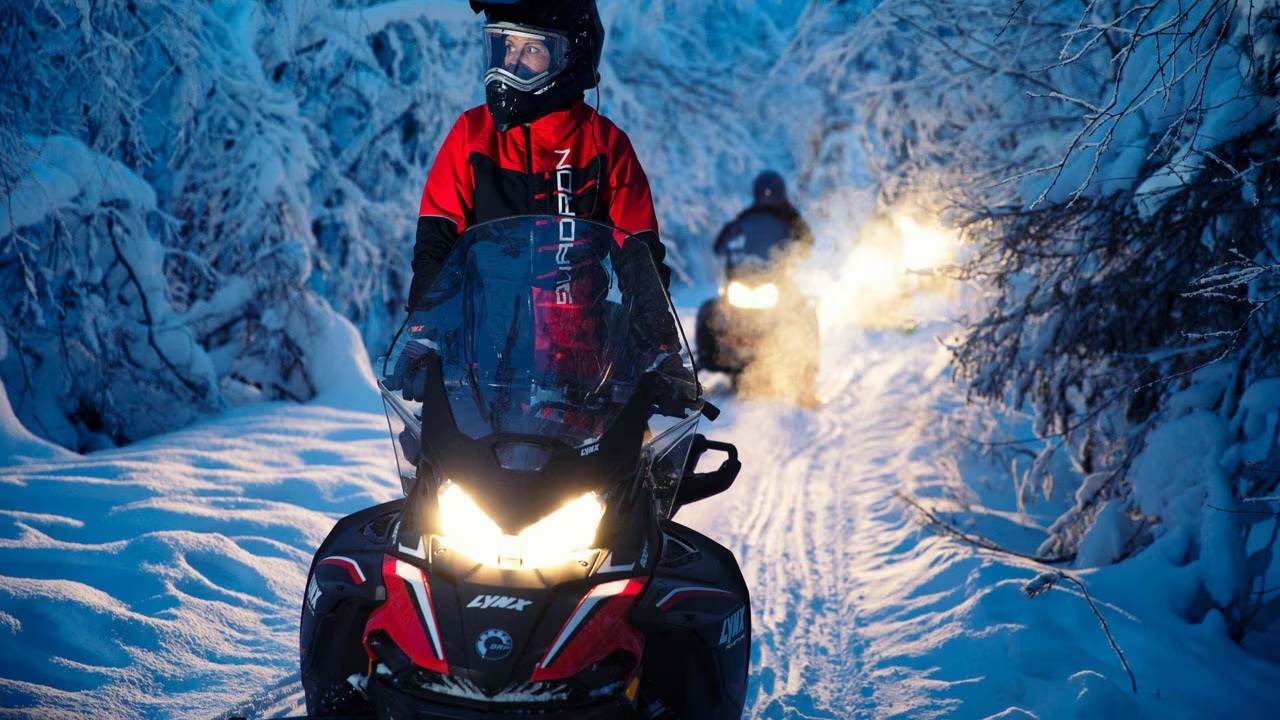 Man standing on a snowmobile, another snowmobile coming from behing, background is snowy forest in Rovaniemi, Lapland, Finland, PolarMax Adventures