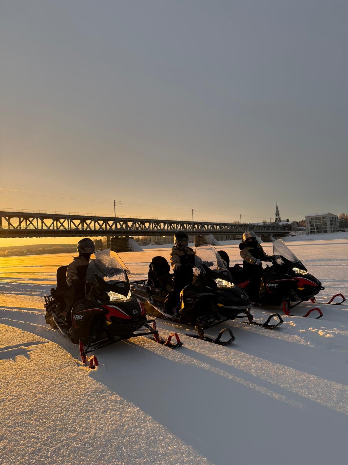 a group of people on snowmobiles in the snow with a railway bridge on the backround, Lapland Royal Adventures, Rovaniemi, Lapland, Finland